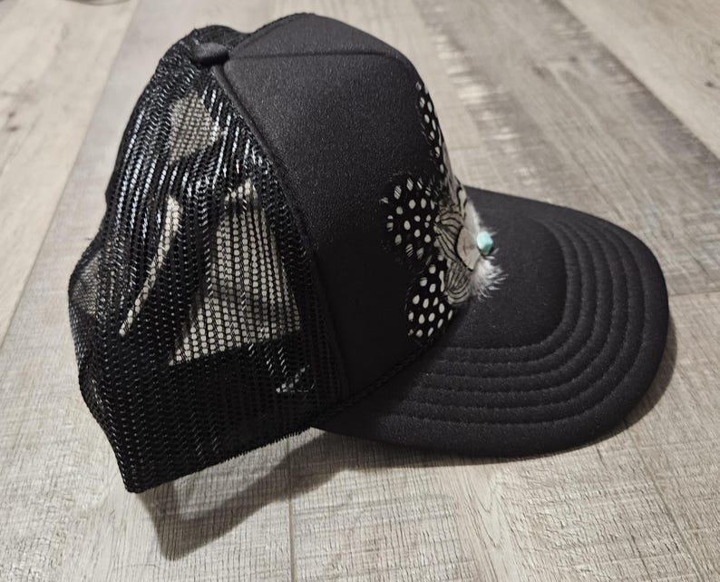 K&ouml;nnte beinhalten: Eine schwarze Mesh-Trucker-Kappe mit einem schwarz-wei&szlig;en Muster und einem t&uuml;rkisfarbenen Feder-Akzent.