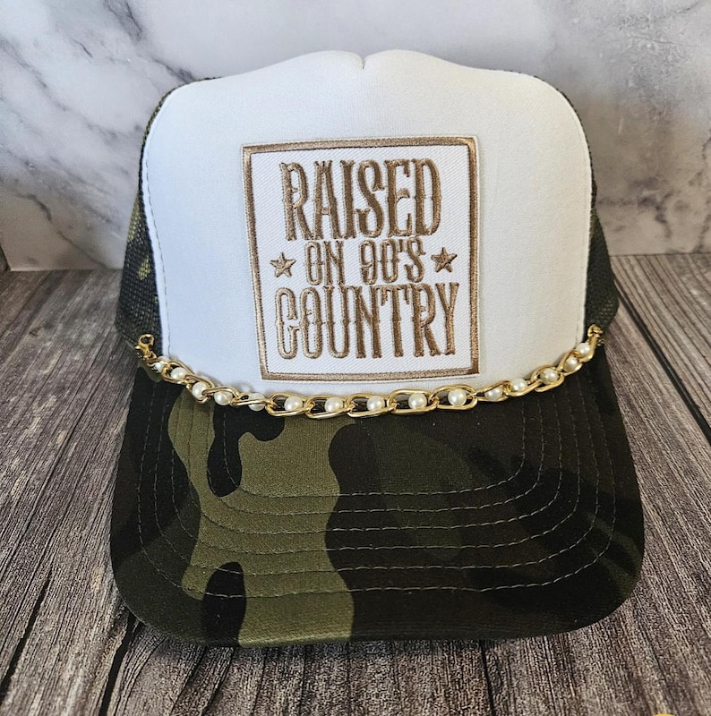 Pode incluir: Bon&eacute; de caminhoneiro com painel frontal branco bordado com "RAISED ON 90'S COUNTRY" em fio dourado. A aba e a parte inferior s&atilde;o camufladas, e uma corrente dourada com p&eacute;rolas adorna a aba. A parte de tr&aacute;s em malha &eacute; preta.
