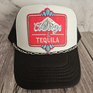 Puede incluir: Un gorro de camionero blanco y negro con un parche rojo y blanco que dice "Cowboys & Tequila". El parche tiene un diseño de montaña azul y una cadena plateada alrededor del borde.