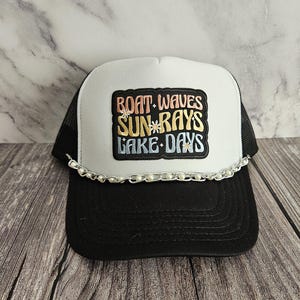 Può includere: Un cappello da camionista bianco e nero con una toppa ricamata colorata che dice "Boat Waves Sun Rays Lake Days". Il cappello ha una catena con perline bianche attorno al bordo.