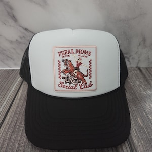 May include: A black and white trucker hat with a white front panel. The front panel features a patch with the words "FERAL MOMS" and "Social Club" with a graphic of a tiger and a person riding it. The hat has a curved brim.