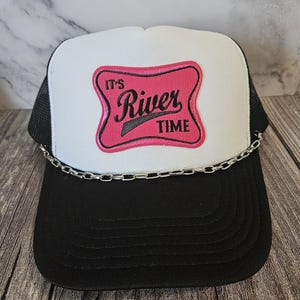 May include: A black and white trucker hat with a curved brim and a mesh back. The front panel is white with a pink and black patch that reads "It's River Time." A silver chain is attached to the brim.