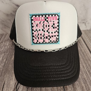 Puede incluir: Gorra de camionero blanca con malla negra en la parte posterior. La gorra tiene un parche a cuadros blanco y negro con el texto "SMALL TOWN SMOKE SHOW" en letras rosas.
