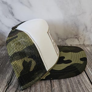 Pode incluir: Bon&eacute; de caminhoneiro camuflado com painel frontal branco e costas em malha. A aba e as costas em malha t&ecirc;m um padr&atilde;o de camuflagem verde e marrom. O bon&eacute; tem um patch marrom com texto.