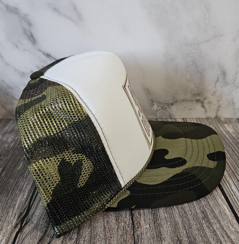 Pode incluir: Bon&eacute; de caminhoneiro camuflado com painel frontal branco e costas em malha. A aba e as costas em malha t&ecirc;m um padr&atilde;o de camuflagem verde e marrom. O bon&eacute; tem um patch marrom com texto.
