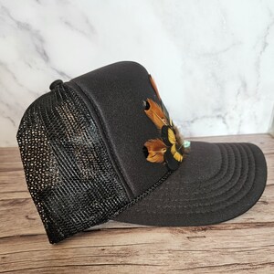 Pode incluir: Bon&eacute; de caminhoneiro preto de malha com um design de penas marrons e amarelas na frente.