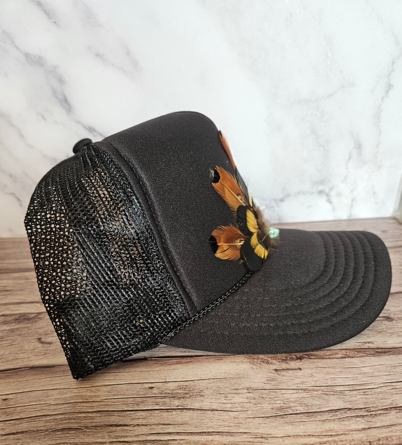 Pode incluir: Bon&eacute; de caminhoneiro preto de malha com um design de penas marrons e amarelas na frente.