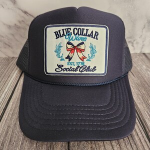 Pode incluir: Boné de caminhoneiro azul marinho com um patch quadrado com o texto "BLUE COLLAR Wives Social Club" e um desenho de laço. O boné tem uma parte traseira de malha e uma aba curva. O texto "EST. 1776" também está no patch.