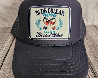 Può includere: Cappellino da camionista blu navy con una toppa quadrata con la scritta "BLUE COLLAR Wives Social Club" e un disegno a fiocco. Il cappellino ha un retro in rete e una visiera curva. Il testo "EST. 1776" è presente anche sulla toppa.