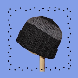 May include: A knitted beanie hat in black and gray. The hat is divided into two sections, with a black lower portion and a gray upper section. The hat is displayed on a wooden stand against a light blue background.