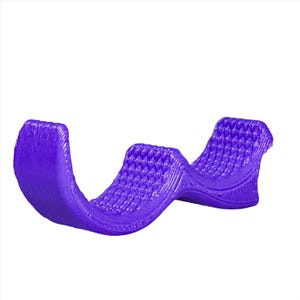 May include: A purple, textured, flexible, plastic support for a phone or tablet. The support is shaped like a wave and has two curves.
