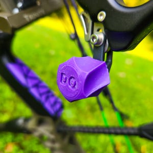 May include: A purple 3D-printed archery bow limb dampener with a hexagonal shape and two circular indentations on the front face. The component is attached to a black metal bow frame with a silver bolt, featuring a textured surface finish.