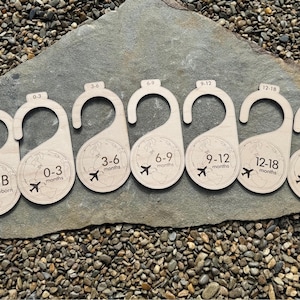 Puede incluir: Conjunto de 8 perchas de madera con un diseño de mapa del mundo y un gráfico de avión. Cada percha está etiquetada con un rango de edad diferente para la ropa de bebé: NB, 0-3 meses, 3-6 meses, 6-9 meses, 9-12 meses, 12-18 meses y 18-24 meses.