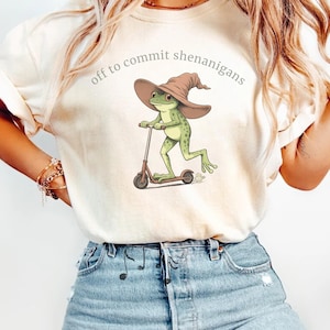 May include: Cream-colored t-shirt featuring a whimsical illustration of a green frog wearing a brown witch hat and riding a brown scooter. The text "off to commit shenanigans" is arched above the frog. The shirt is made of soft material.
