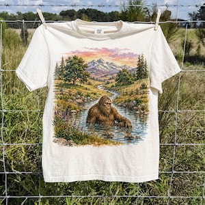 Puede incluir: Una camiseta de manga corta color crema con un estampado gráfico de una criatura marrón y peluda que se asemeja a Bigfoot vadeando un arroyo. La escena incluye un paisaje con árboles verdes, un río serpenteante y montañas nevadas al atardecer.