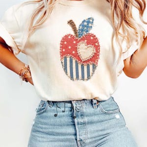 Puede incluir: Una camiseta de color crema con un gráfico central de una manzana compuesto por varios patrones de tela. El diseño de la manzana incluye lunares rojos, rayas azules y blancas, una hoja de cuadros vichy azules y un corazón central color crema.