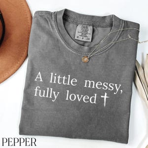 Puede incluir: Una camiseta gris con el texto blanco "A little messy, fully loved". Un collar dorado con un colgante está sobre la camiseta. La camiseta está etiquetada como "Comfort Colors" y es talla S-P-CH.