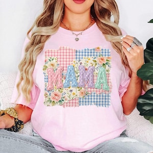 Puede incluir: Una camiseta rosa claro con la palabra "MAMA" en colores pastel. Las letras están sobre un diseño de patchwork con estampados florales y de cuadros. Una prenda casual para el día a día.