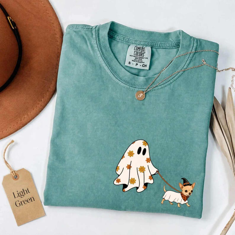 May include: A seafoam green crewneck t-shirt featuring a graphic of a white ghost with orange flower patterns walking a small tan dog wearing a black witch hat and a red plaid bandana. The shirt has a Comfort Colors label inside the collar.