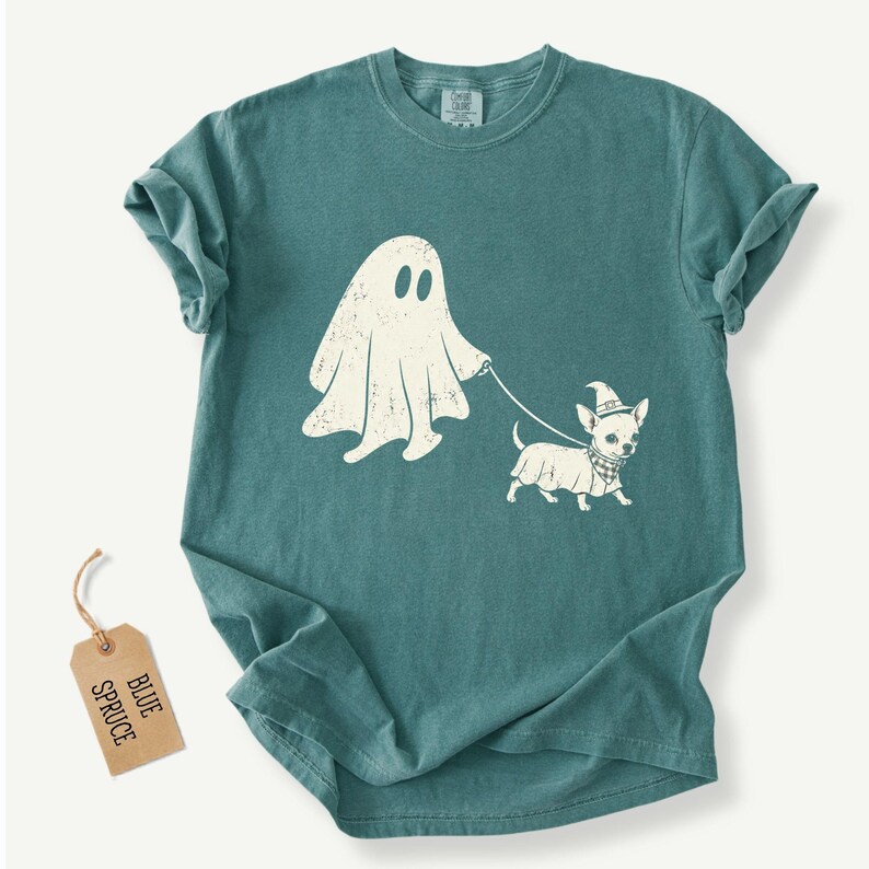 May include: A teal short-sleeve t-shirt featuring a white graphic of a ghost walking a small dog wearing a witch hat and a checkered bandana. The ghost holds a leash attached to the dog. A tan paper tag labeled BLUE SPRUCE is attached.