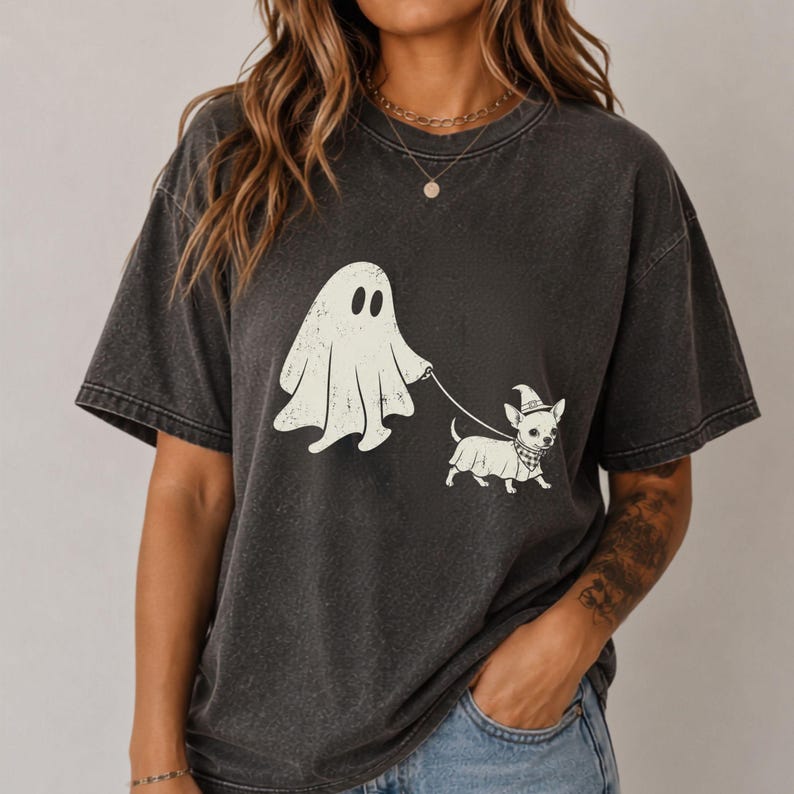 May include: A charcoal gray mineral-washed t-shirt featuring a graphic print of a white ghost walking a small dog. The dog is wearing a witch hat and a checkered bandana. The ghost holds a thin leash connected to the dog's collar in this design.
