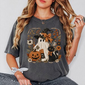 Puede incluir: Una camiseta gris carbón de manga corta con un estampado gráfico de un fantasma con sombrero de bruja y lazo naranja, una calabaza de Halloween y un gato negro con lazo naranja, rodeados de flores y follaje de temática otoñal.