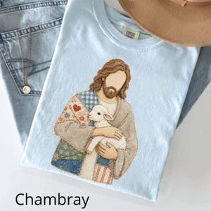 Puede incluir: Una camiseta azul claro con un estampado gráfico de una figura con cabello largo castaño y barba, que viste una túnica estilo patchwork y sostiene un pequeño cordero blanco. El diseño incluye patrones florales, de cuadros y rayas en tonos tierra.