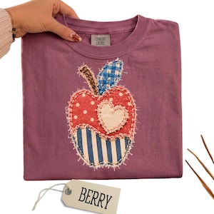 May include: A berry-coloured short-sleeve t-shirt featuring a central apple-shaped fabric appliqué. The apple design is composed of red polka-dot, blue-and-white striped, and blue gingham fabric patches with a central cream-coloured heart.