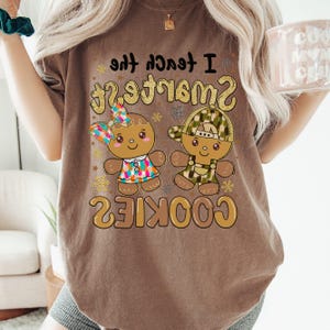 Puede incluir: Una camiseta marrón de manga corta con un gráfico de dos personajes de pan de jengibre y el texto "I teach the SMARTEST COOKIES". Un personaje lleva un lazo y un vestido de colores, mientras que el otro lleva una gorra y un peto de camuflaje.