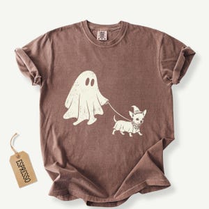 May include: A brown short-sleeve t-shirt featuring a cream-colored graphic of a ghost walking a small dog wearing a witch hat and a checkered bandana on a leash. A rectangular tag attached to the shirt displays the word ESPRESSO in black text.