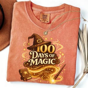 Puede incluir: Camiseta de color coral con un gráfico de un sombrero de mago, el texto "100 Days of Magic", un libro de hechizos y una varita. La camiseta tiene cuello redondo y una etiqueta Comfort Colors.