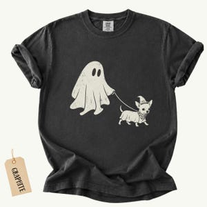 May include: A charcoal gray short-sleeve t-shirt featuring a distressed cream-colored graphic of a ghost walking a small dog wearing a witch hat and a checkered bandana. A rectangular tag labeled GRAPHITE is attached to the bottom left corner.