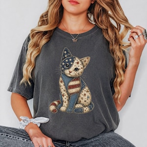 May include: Dark gray t-shirt featuring a patriotic cat design. The cat is depicted with a patchwork pattern incorporating the American flag, including stars and stripes. The cat is sitting, with a detailed and colorful design.