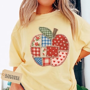 Puede incluir: Una camiseta de color amarillo pálido con un gráfico de manzana grande compuesto por varios parches de tela, incluyendo flores rojas, cuadros azules, cuadros rojos y blanco con corazones rojos. Una etiqueta muestra el texto BANANNA.
