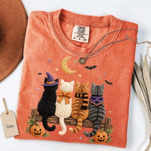 May include: An orange short-sleeve t-shirt featuring a graphic of four cats sitting on a wooden fence. The cats wear a witch hat, a bow, a scarf, and a bandana. The design includes a crescent moon, stars, bats, and two jack-o'-lanterns. The shirt has a Comfort Colors brand label.
