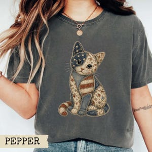 May include: A charcoal grey short-sleeve t-shirt featuring a graphic of a sitting cat. The cat has a patchwork design incorporating stars and stripes in red, white, and blue, along with floral patterns in muted earth tones across its body.