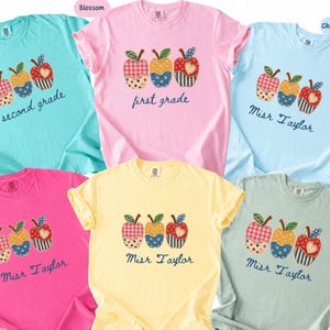 May include: Six short-sleeved crew neck t-shirts in mint, blossom, chambray, crunchberry, yellow, and bay colors. Each shirt features a graphic of three apples with varied fabric-style patterns and text reading second grade, first grade, or Misr Taylor.