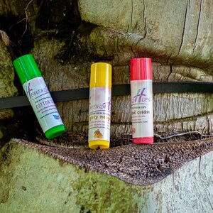 May include: Three lip balm tubes in green, yellow, and red, each with a white label and text. The green tube reads "Cotton Candy," the yellow tube says "Mighty Mint," and the red tube says "Hand Cream." The tubes are displayed on a textured, brown surface.