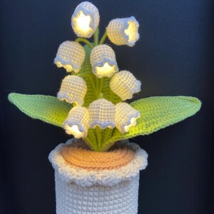 May include: A crocheted lily of the valley lamp with illuminated white flower heads and green leaves. The plant sits in a white pot with a scalloped edge. The lamp is a decorative home accent.