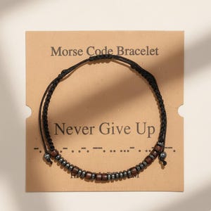 May include: A black cord Morse code bracelet with brown and silver beads, displayed on a tan card. The card reads "Morse Code Bracelet" and "Never Give Up". The bracelet is a stylish accessory.
