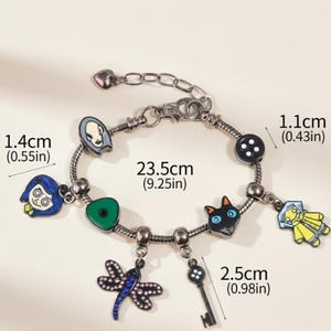 May include: A silver-toned charm bracelet featuring colorful enamel charms. The bracelet includes charms of a girl, a cat, a key, a dragonfly, a button, and a character with a pale face. The bracelet is 9.25 inches long.