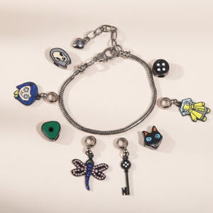 May include: A silver-toned charm bracelet with multiple dangling charms. Charms include a black button, a cat face, a key, a dragonfly, a green triangle, and character-themed charms in blue, yellow, and gray.