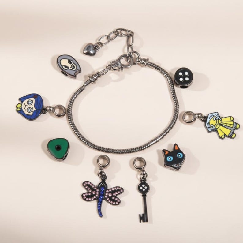 May include: A silver-toned charm bracelet with multiple dangling charms. Charms include a black button, a cat face, a key, a dragonfly, a green triangle, and character-themed charms in blue, yellow, and gray.