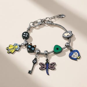 May include: Silver charm bracelet featuring multiple colorful charms. Charms include a black cat, a key, a dragonfly, a button, and character faces. The bracelet is on a light beige background.
