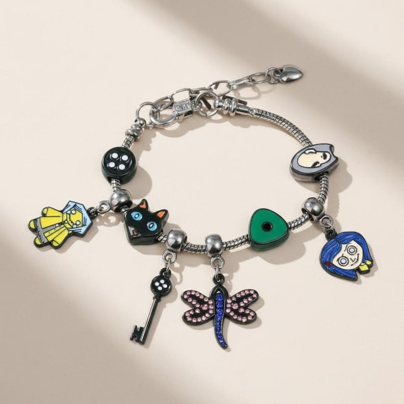 May include: Silver charm bracelet featuring multiple colorful charms. Charms include a black cat, a key, a dragonfly, a button, and character faces. The bracelet is on a light beige background.