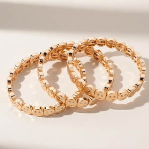 May include: Three gold-colored bracelets with a repeating smiley face design. Each bracelet is made of small, rounded links, with each link featuring a smiling face and small, clear accents. The bracelets are arranged on a white surface.