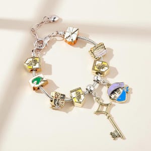 May include: Silver-toned charm bracelet featuring colorful enamel charms with various shield and emblem designs. A gold-colored key charm hangs from the bracelet. The charms are in shades of orange, yellow, green, black, and blue.