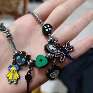 May include: A silver charm bracelet featuring several themed charms. Charms include a black cat, a key, a green button, and characters from a movie. The bracelet has a black button charm and a dragonfly charm.