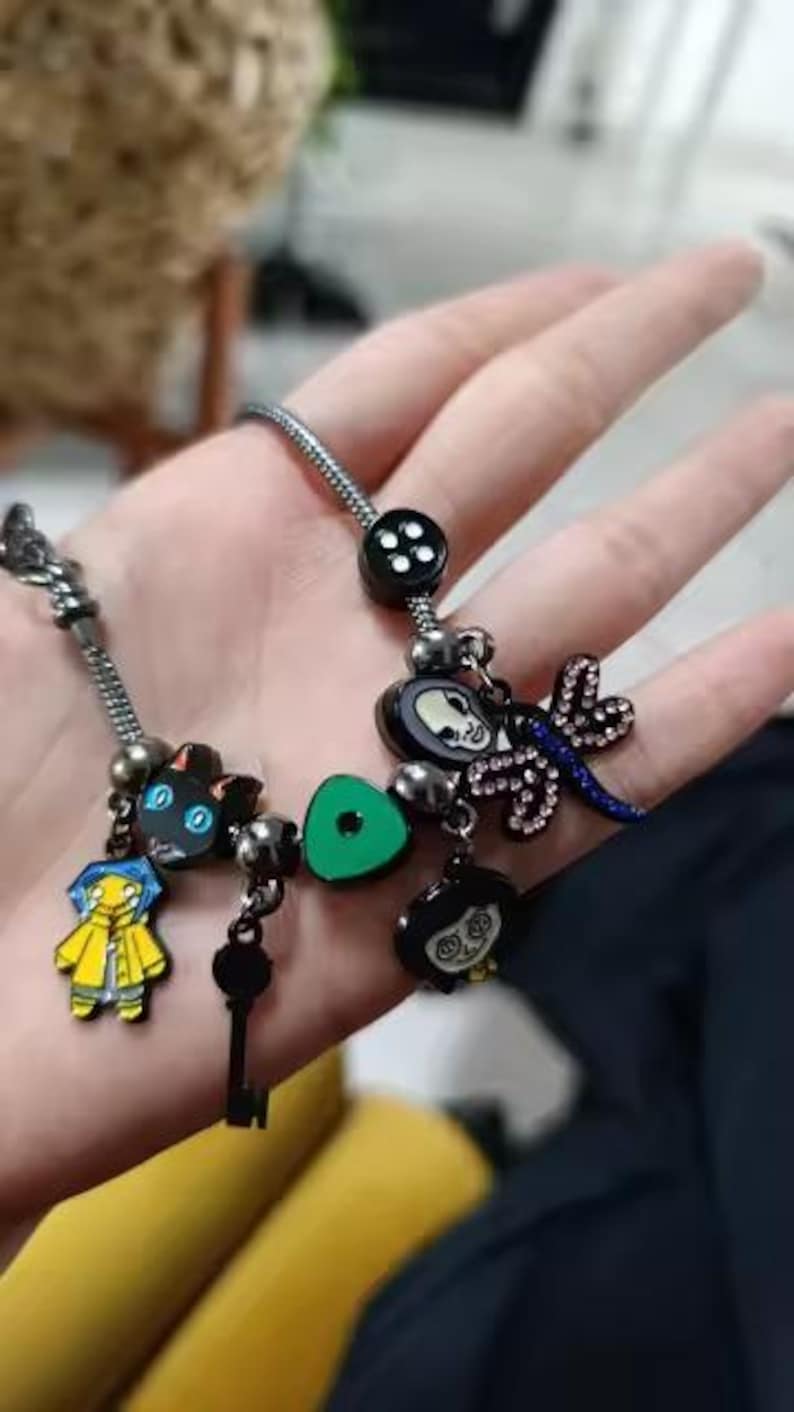 May include: A silver charm bracelet featuring several themed charms. Charms include a black cat, a key, a green button, and characters from a movie. The bracelet has a black button charm and a dragonfly charm.