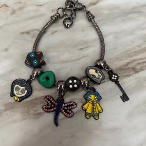May include: Silver charm bracelet featuring various character charms. Charms include a black cat, a green button, a dragonfly, a key, and character faces. The bracelet has a lobster clasp closure.